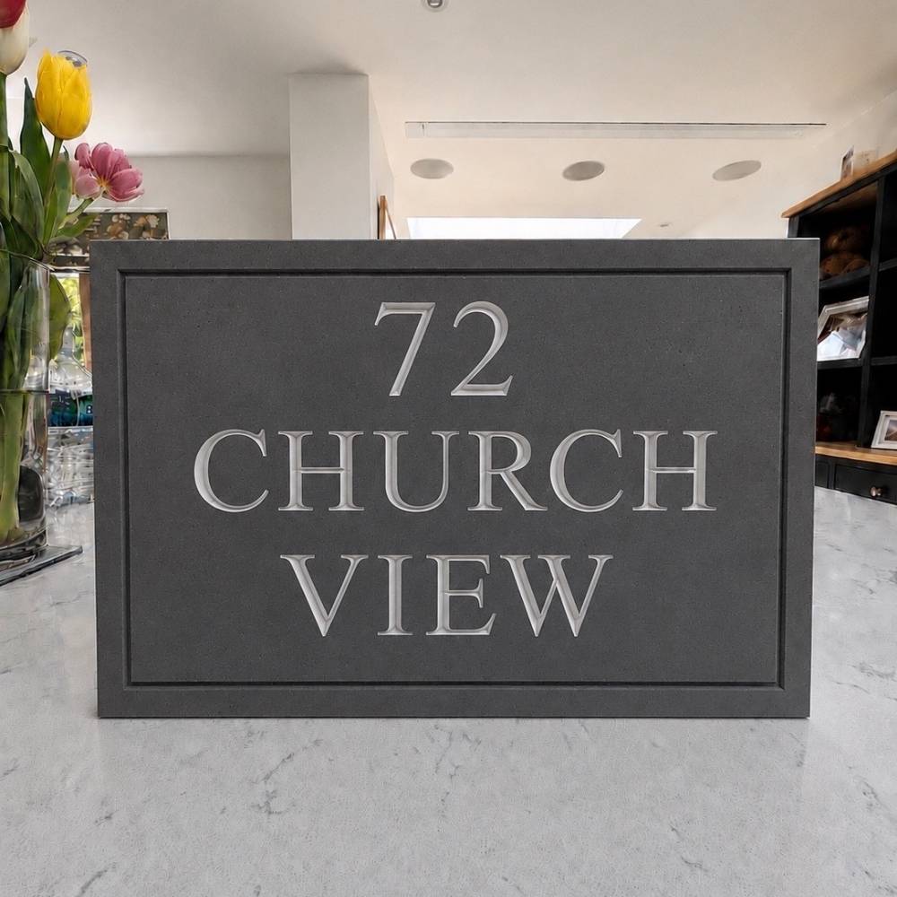 A rectangular dark slate house number plaque with raised silver lettering reading '72 CHURCH VIEW' on a light marble surface, with a blurred indoor background and flowers at the left edge.