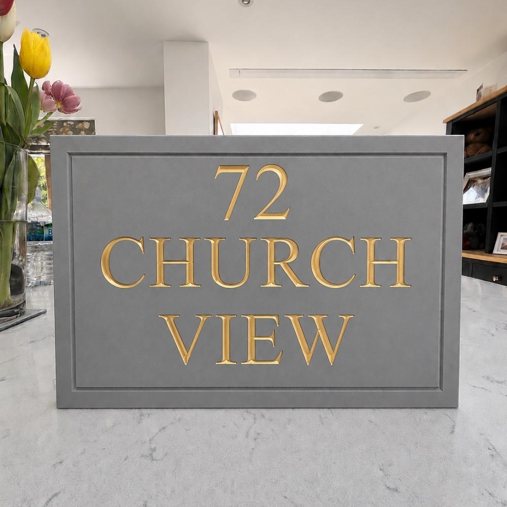 A rectangular grey house number plaque with raised gold lettering reading '72 CHURCH VIEW' on a light marble countertop in a modern interior setting.