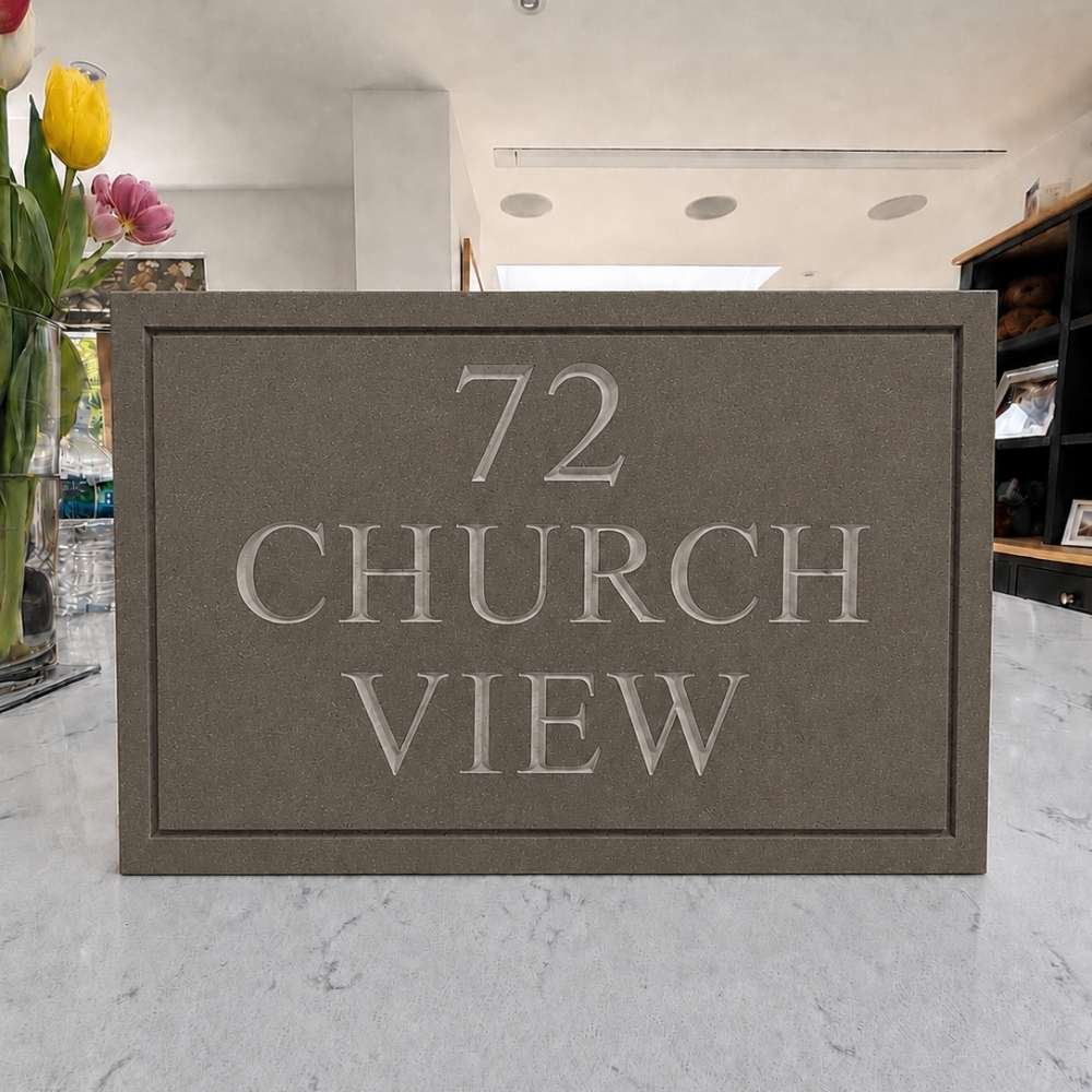 A rectangular mink house plaque with raised lettering reading '72 CHURCH VIEW' sits on a light marble countertop in a store interior. Tulips in a vase and shelving are visible in the background.