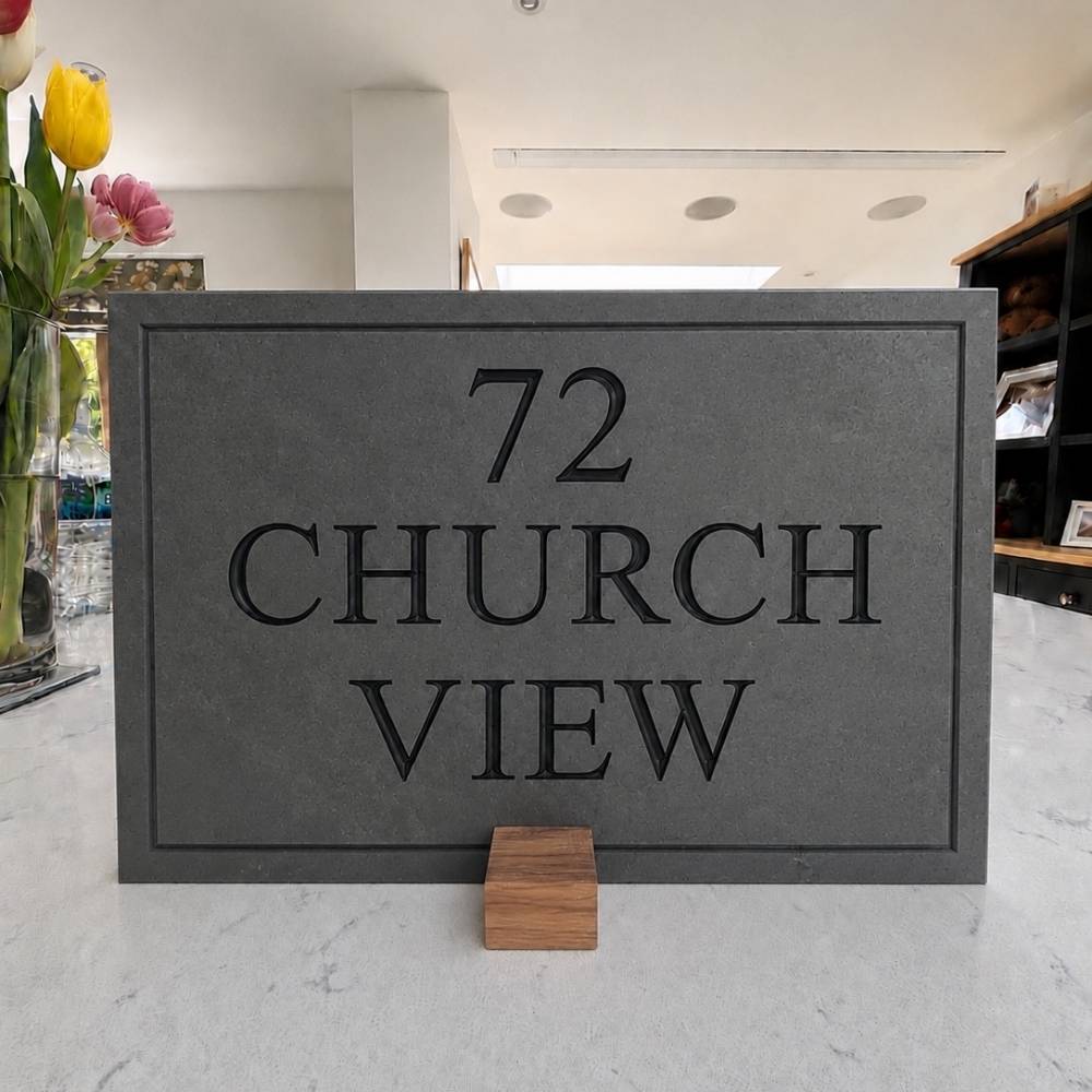 A rectangular slate stone or concrete house number plaque reading '72 CHURCH VIEW,' displayed on a light marble-like surface with a small wooden stand. A real indoor environment is visible in the background, including flowers, shelving, and ceiling lights, so this is Lifestyle rather than Product/Packaging.