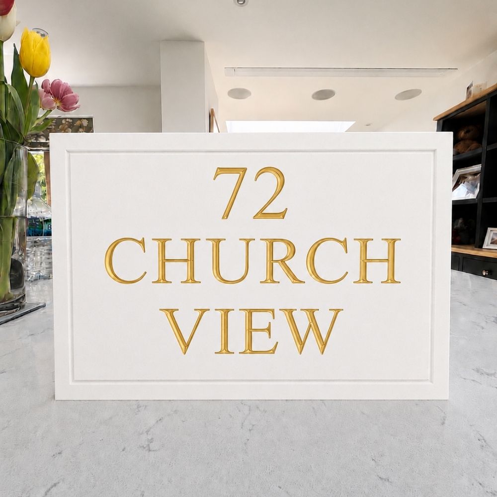 A white rectangular plaque with raised gold lettering reading '72 CHURCH VIEW' on a light marble surface, with flowers and an indoor room visible in the background.