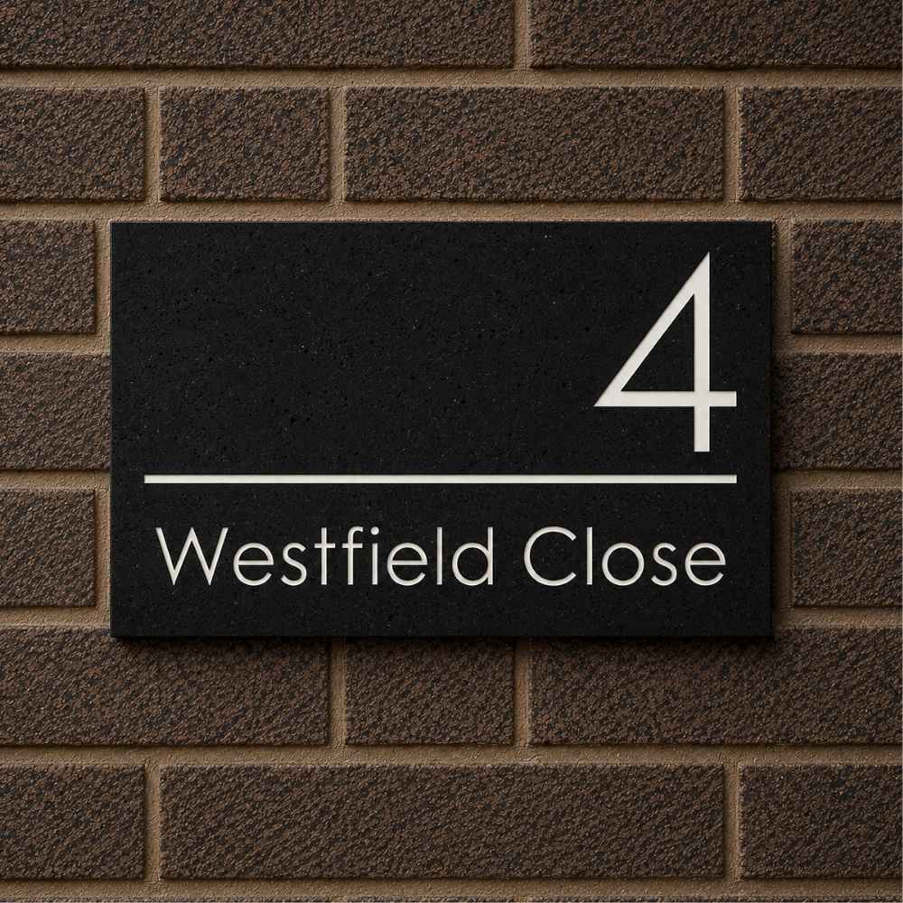 A rectangular charcoal house number sign mounted on a brown brick wall, with white text reading '4 Westfield Close'.