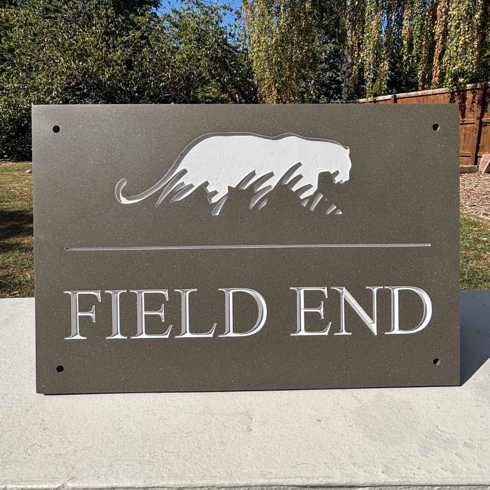A rectangular mink entrance sign and off white engraved lettering reading FIELD END, plus a silver animal silhouette near the top. The sign is photographed outdoors on a concrete surface with trees and a fence in the background.