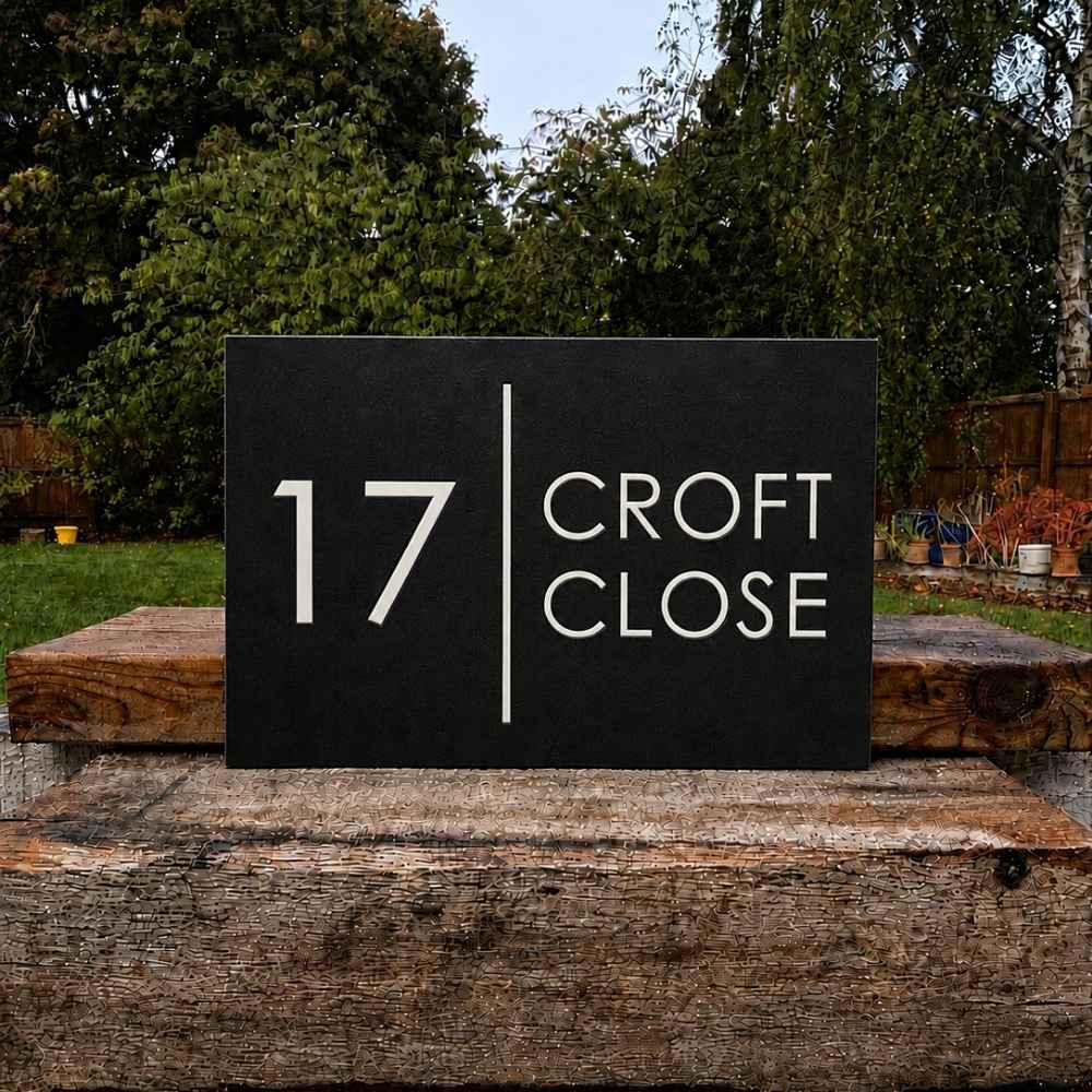 A rectangular black house number plaque reading '17 CROFT CLOSE' in white lettering, displayed outdoors on a rustic wooden post with trees and a garden background.