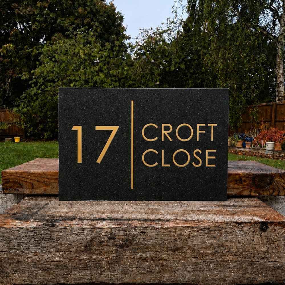 A rectangular charcoal house number plaque with gold lettering reading '17 CROFT CLOSE', displayed outdoors on a rustic wooden wall with trees and garden fencing in the background.