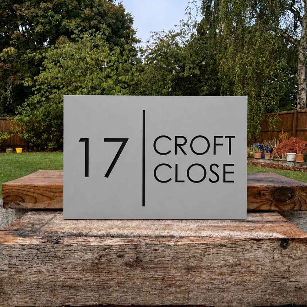 A rectangular gray house number sign reading “17 CROFT CLOSE” with a vertical divider, displayed outdoors on a rustic wooden post in a garden setting.