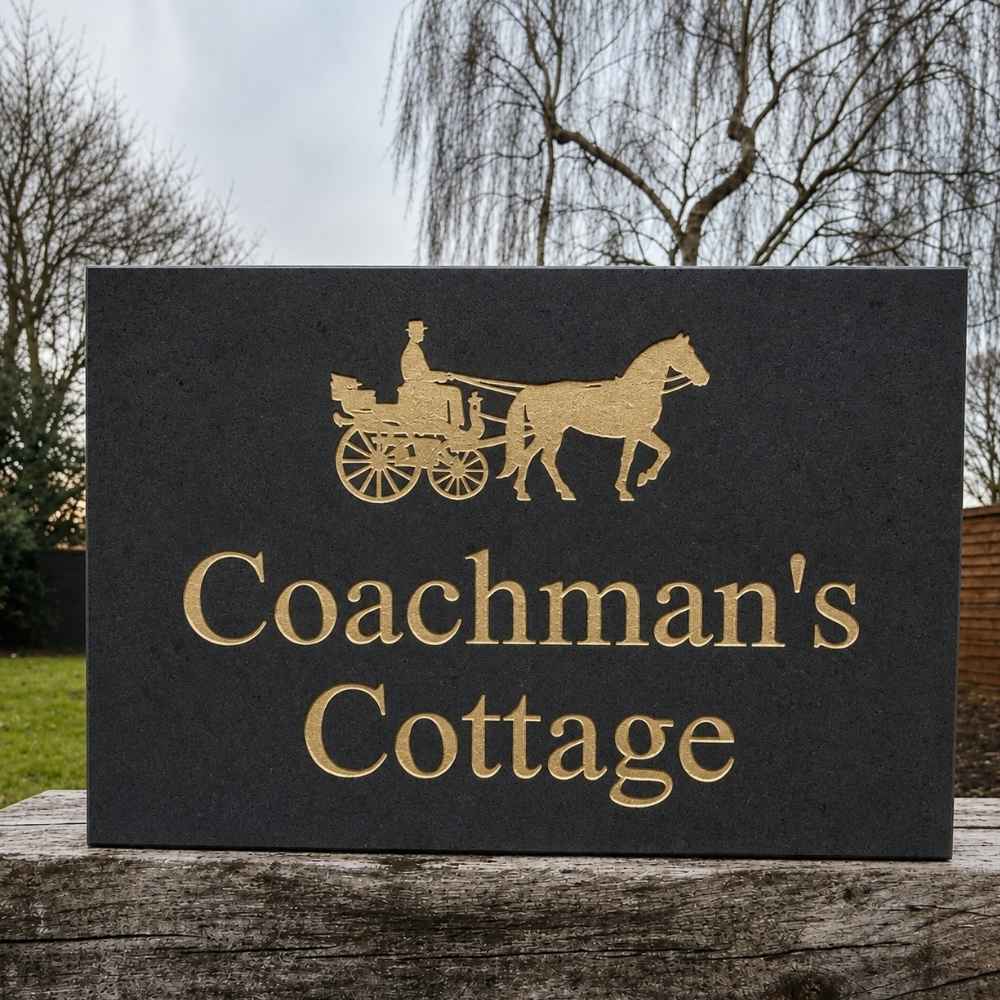 A rectangular black plaque with gold lettering reading 'Coachman's Cottage' and a gold horse-and-buggy silhouette, displayed outdoors on a wooden surface with trees and sky in the background.