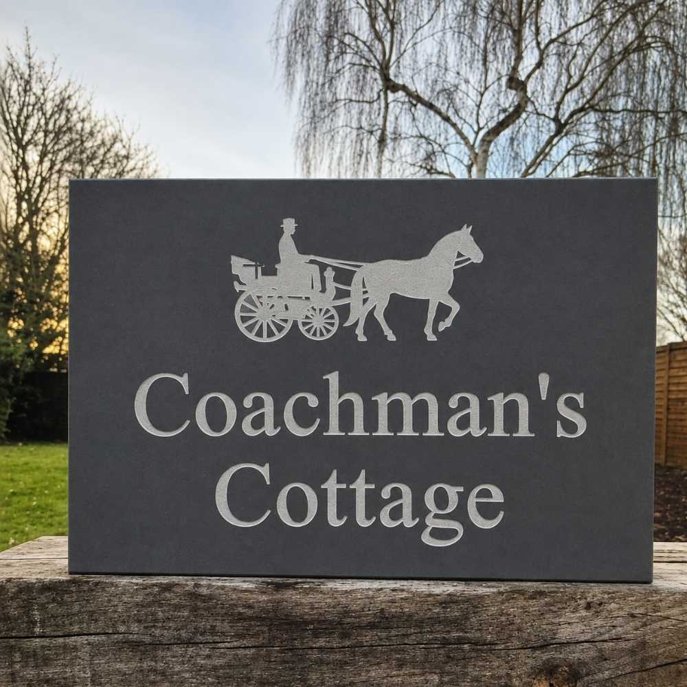 A rectangular dark slate sign with a silver silhouette of a horse-drawn carriage and the words 'Coachman's Cottage,' displayed outdoors on a wooden post with trees and grass in the background.
