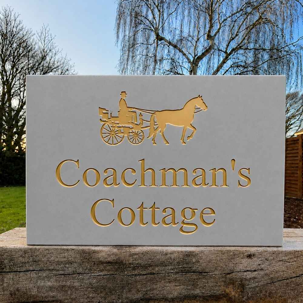 A rectangular grey stone or concrete sign with raised gold lettering reading 'Coachman's Cottage' and a gold horse-and-carriage emblem at the top, photographed outdoors on a wooden post with trees and sky in the background.