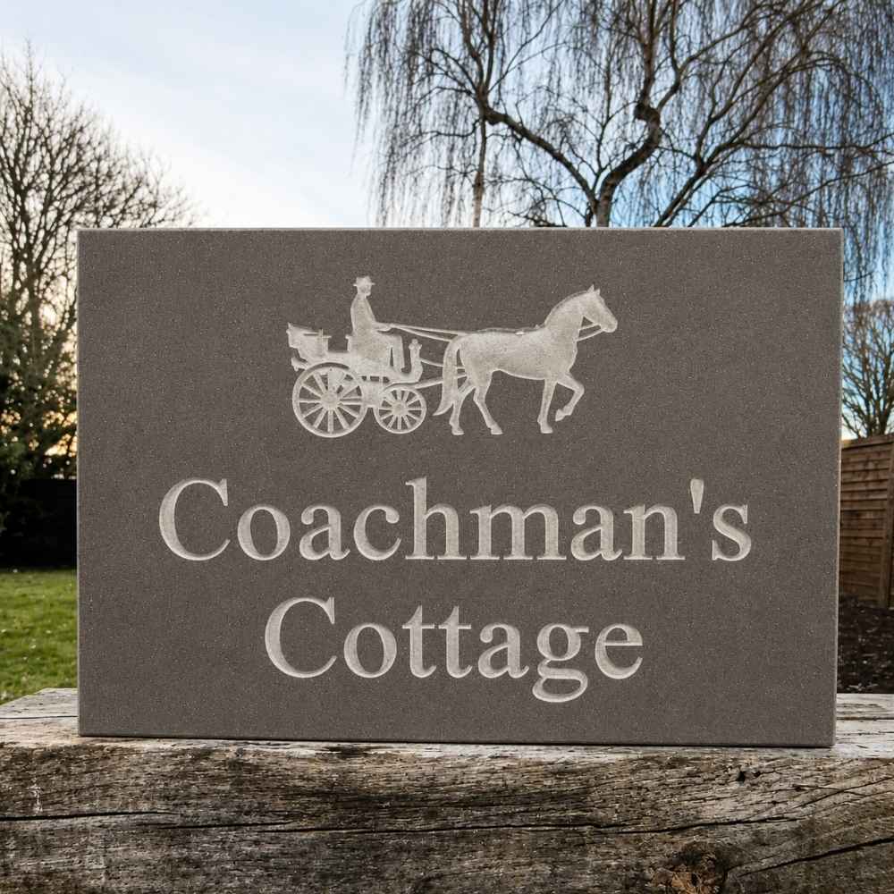 A rectangular mink gray stone plaque engraved with a white coachman and horse silhouette and the words 'Coachman's Cottage,' displayed outdoors on a weathered wooden post with trees and sky in the background.