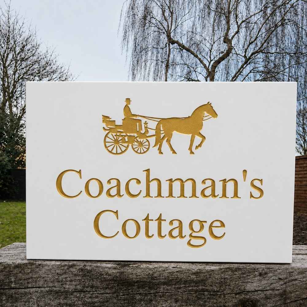 A white rectangular sign with gold lettering reading 'Coachman's Cottage' and a gold silhouette of a horse-drawn carriage, photographed outdoors on a wooden surface with trees and sky in the background.
