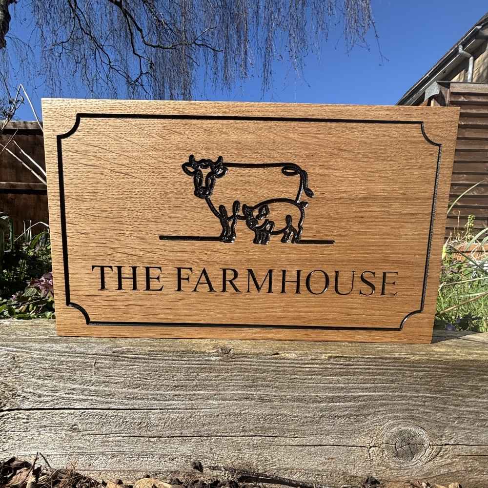 A rectangular oak wooden sign featuring a black engraved illustration of a cow and calf and the text 'THE FARMHOUSE'. The sign is displayed outdoors on a weathered wooden post with a blue sky and garden background.