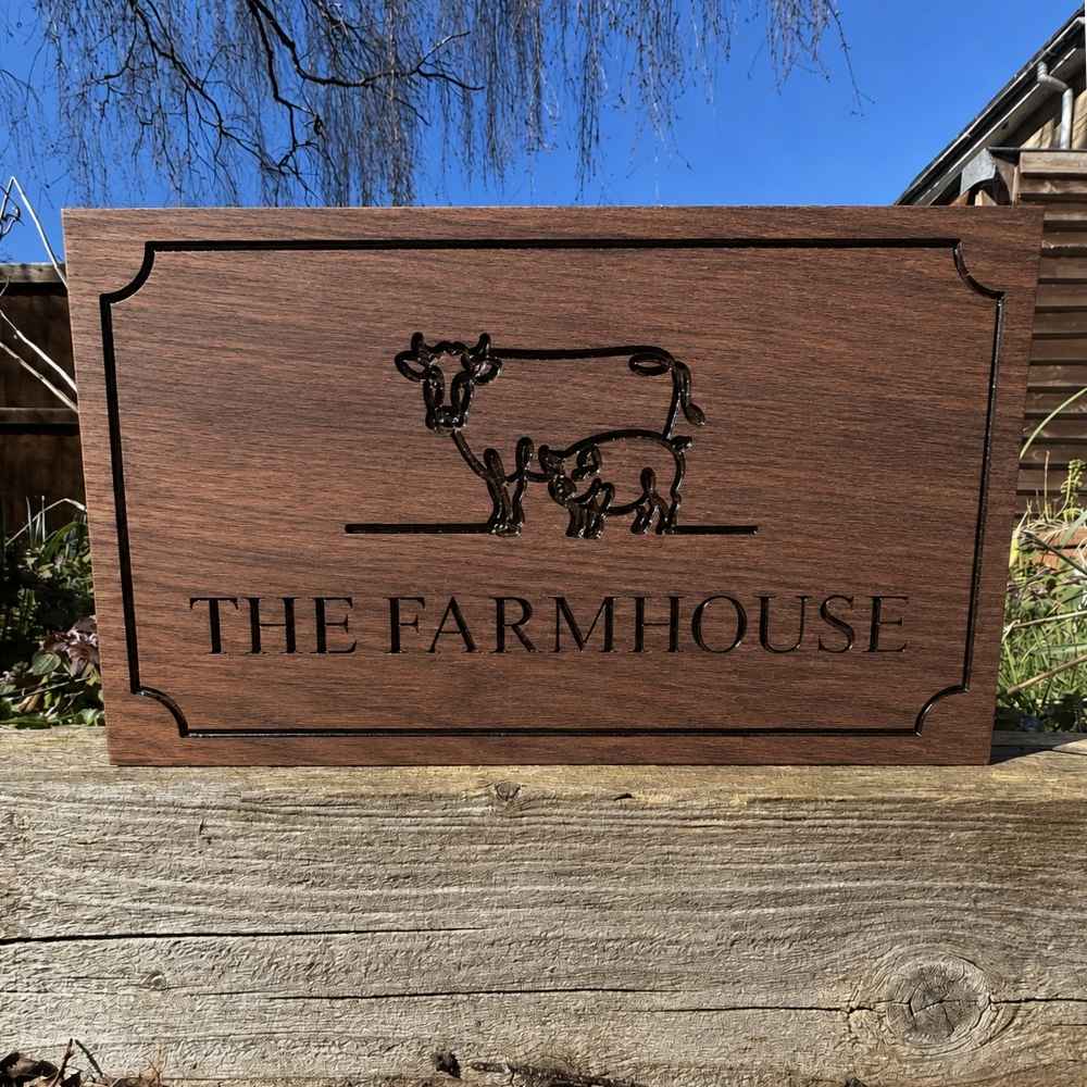 A rectangular walnut wooden sign featuring an engraved silhouette of a cow and calf and the text 'THE FARMHOUSE' in black. The sign is displayed outdoors on a rustic wooden post with a blue sky and tree branches in the background.