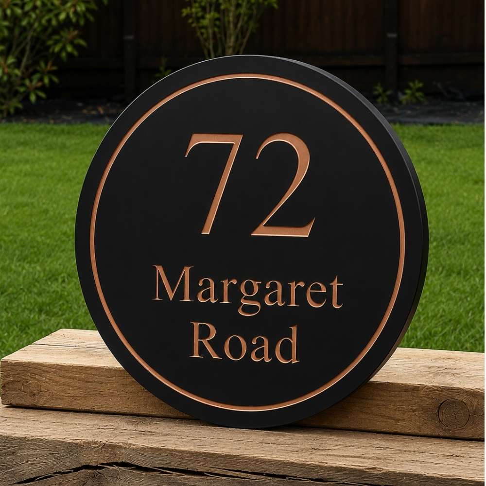 A round charcoal house number plaque with rose gold lettering and border, displaying '72 Margaret Road', resting on a wooden post in an outdoor garden setting.