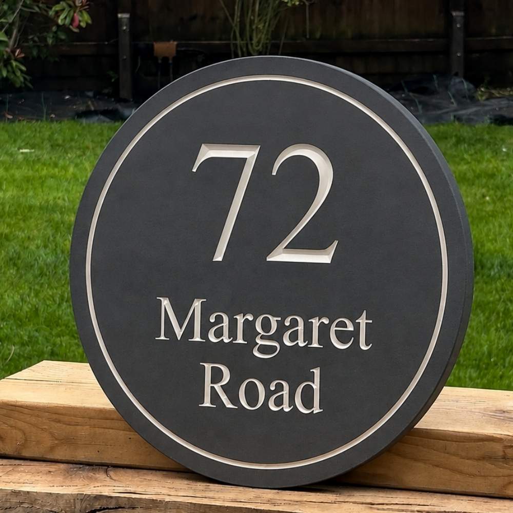 A round dark slate house number sign with silver lettering reading '72 Margaret Road' displayed on a wooden post in a backyard setting with green grass behind it.