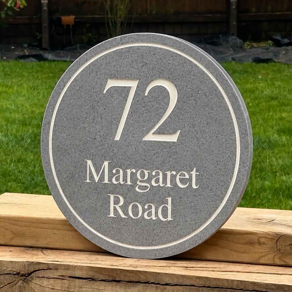 A round charcoal stone house number plaque reading '72 Margaret Road' in white lettering, displayed outdoors on a wooden fence with green grass in the background.