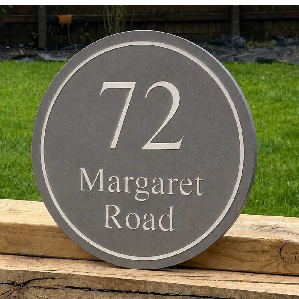 A round grey house number sign reading '72 Margaret Road' with a white border, photographed outdoors on a wooden fence with green grass in the background.