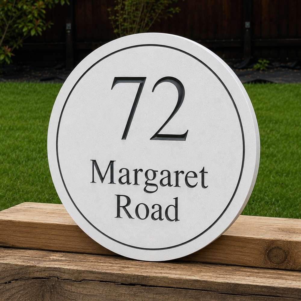 A round white house number plaque with black text reading '72 Margaret Road' resting on a wooden post outdoors with green grass and a dark fence in the background.