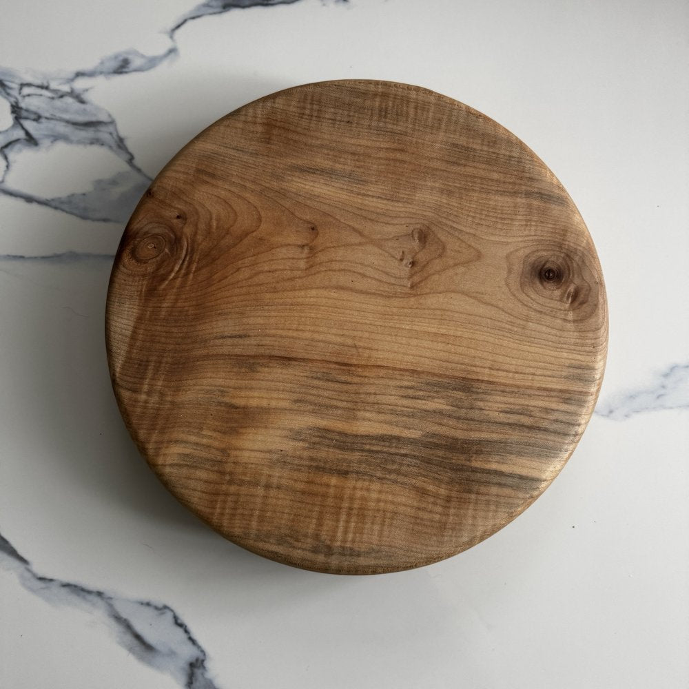 A round wooden cutting board with a natural grain pattern, photographed from directly above on a white marble surface.