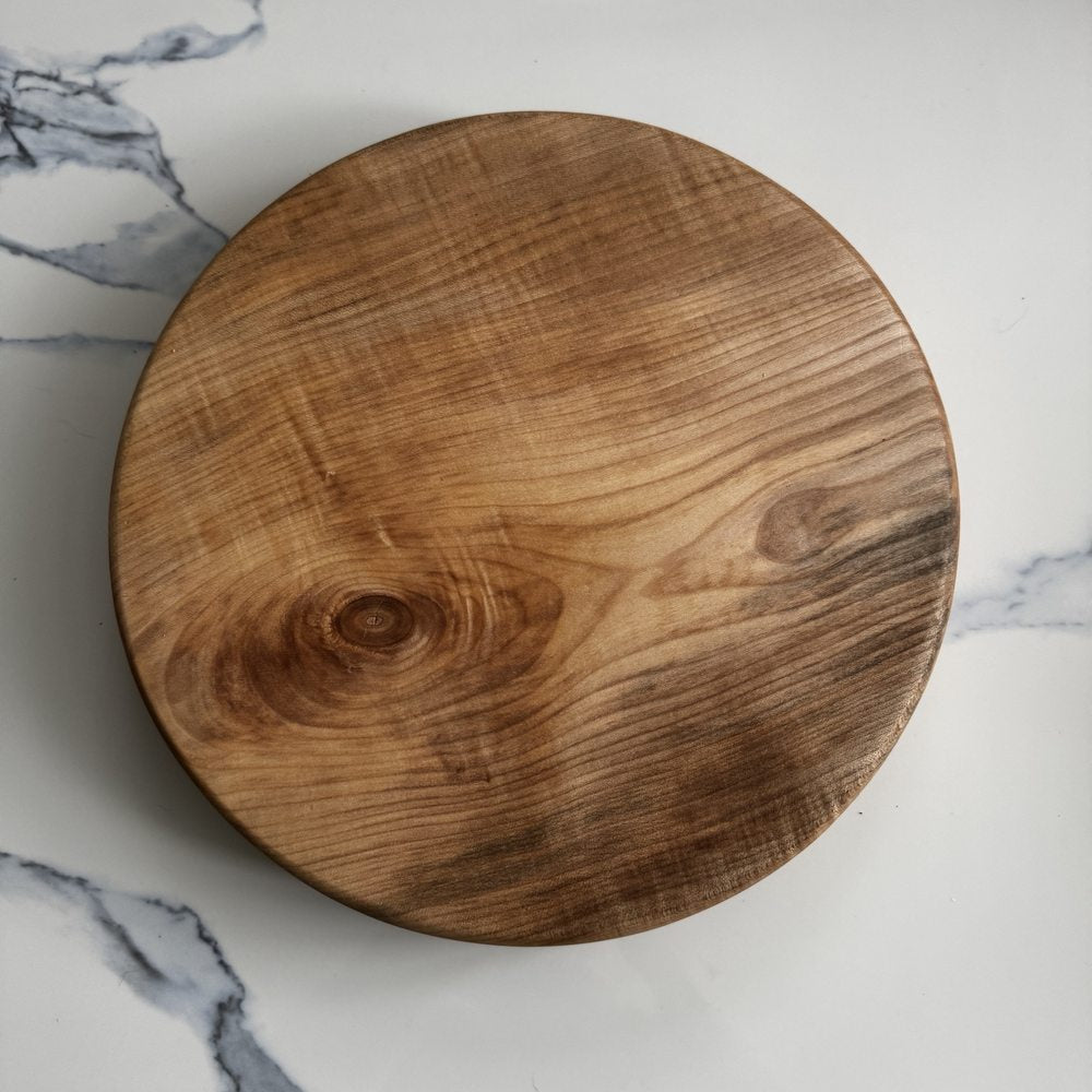 A round wooden cutting board with a prominent natural grain pattern and a single visible knot, photographed from directly above on a white marble surface.