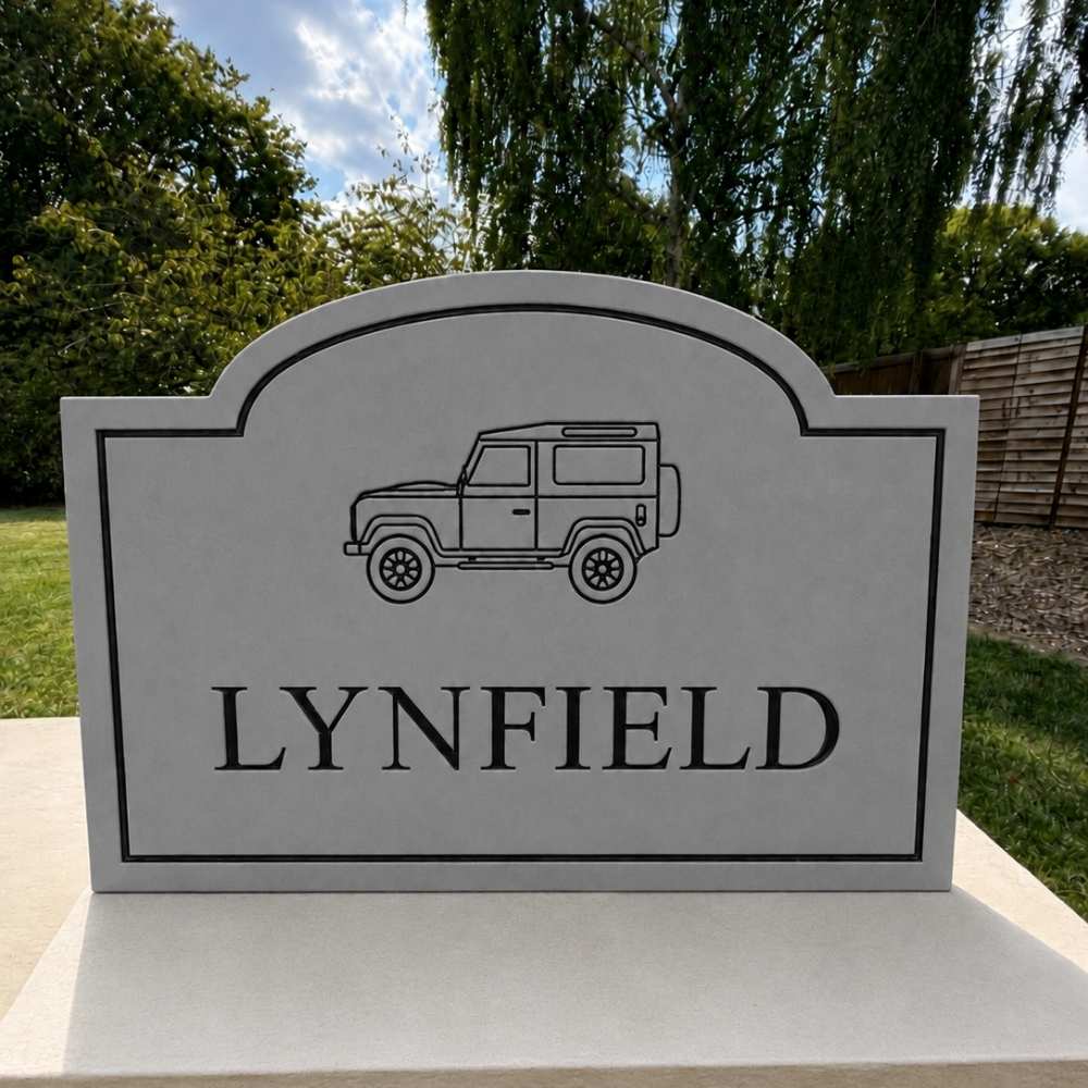 A custom grey rectangular outdoor sign with an arched top, featuring a black line drawing of a classic off-road vehicle and the name 'LYNFIELD' in bold black letters. The sign is mounted on a light-colored stone or concrete base outdoors, with green trees and grass in the background.