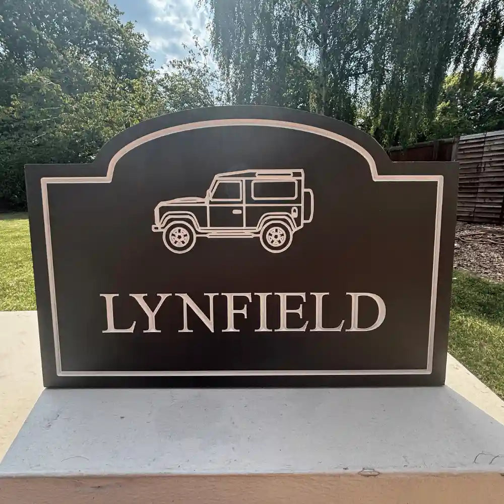 A black sign featuring an engraved off-road vehicle silhouette and the name 'LYNFIELD' in raised letters, mounted on a light-colored stone pillar outdoors.
