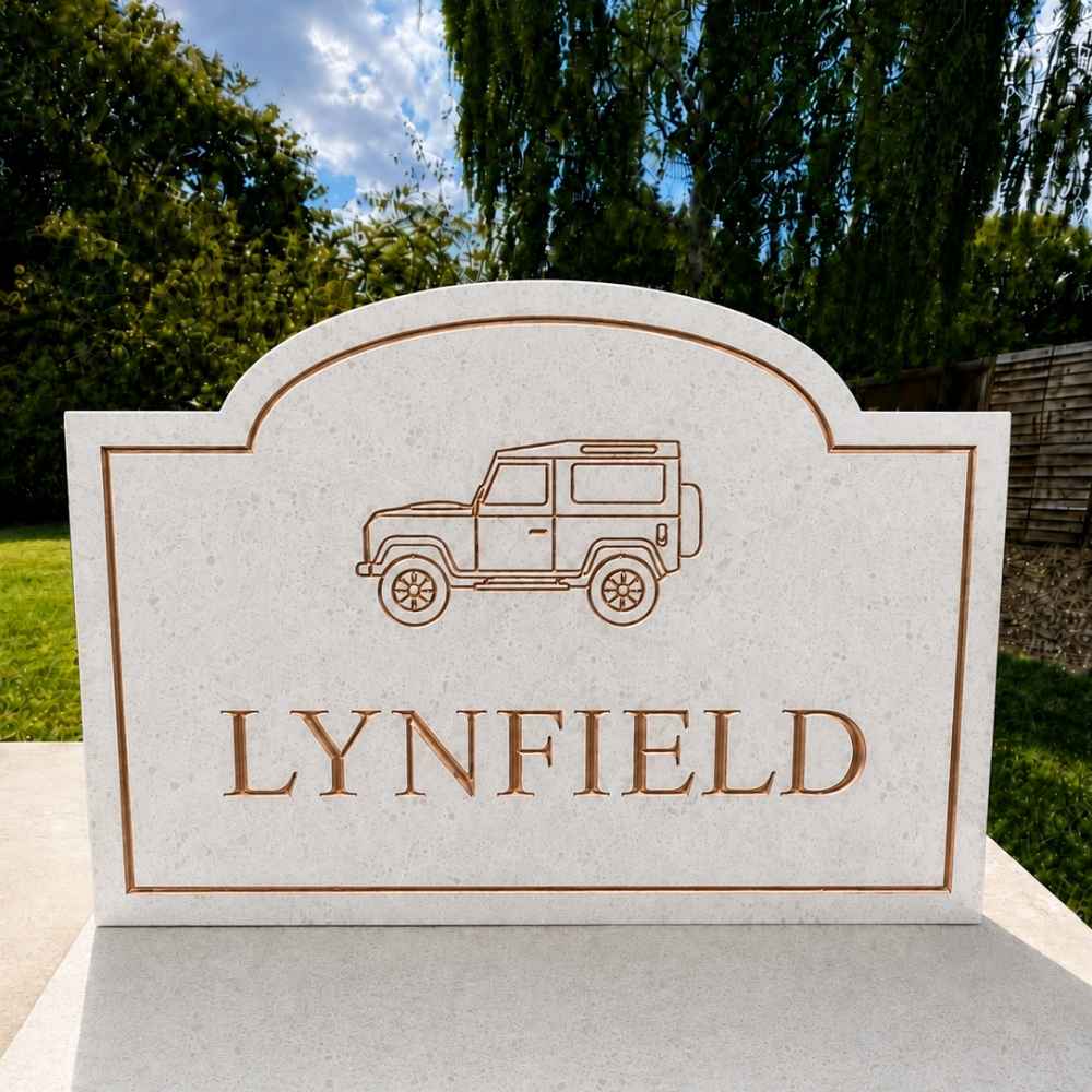 A custom engraved stone quartz sign  featuring a copper outline drawing of a vintage off-road vehicle and the name 'LYNFIELD' in raised brown lettering, set outdoors against a background of green trees and blue sky.