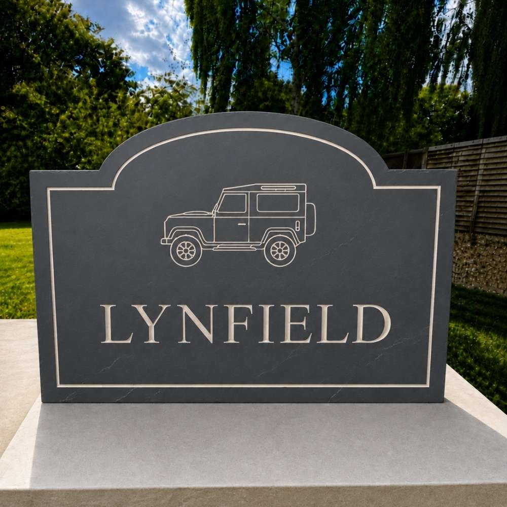 A custom engraved slate sign featuring a white line drawing of a Land Rover and the name 'LYNFIELD' in raised letters, mounted on a light gray stone base with a garden background.