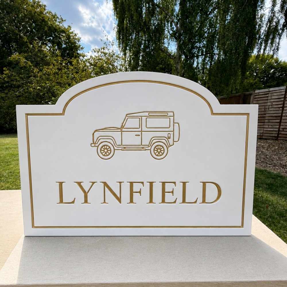 A custom white stone sign with a gold border, featuring an engraved line drawing of a classic Land Rover and the name 'LYNFIELD' in gold capital letters. The sign is displayed outdoors on a stone pedestal with a garden background.