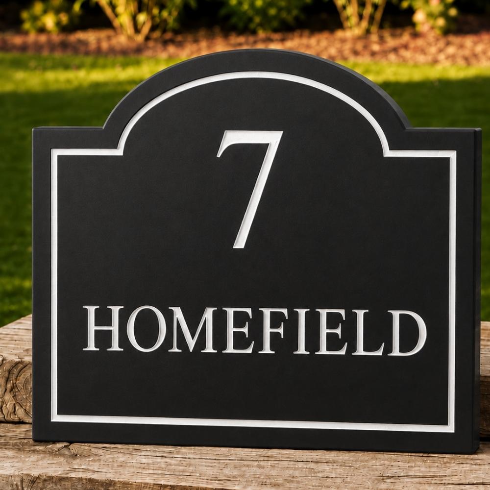 A black arched house number sign with silver lettering that reads '7 HOMEFIELD', displayed outdoors on a wooden post with a blurred green lawn and bushes in the background.