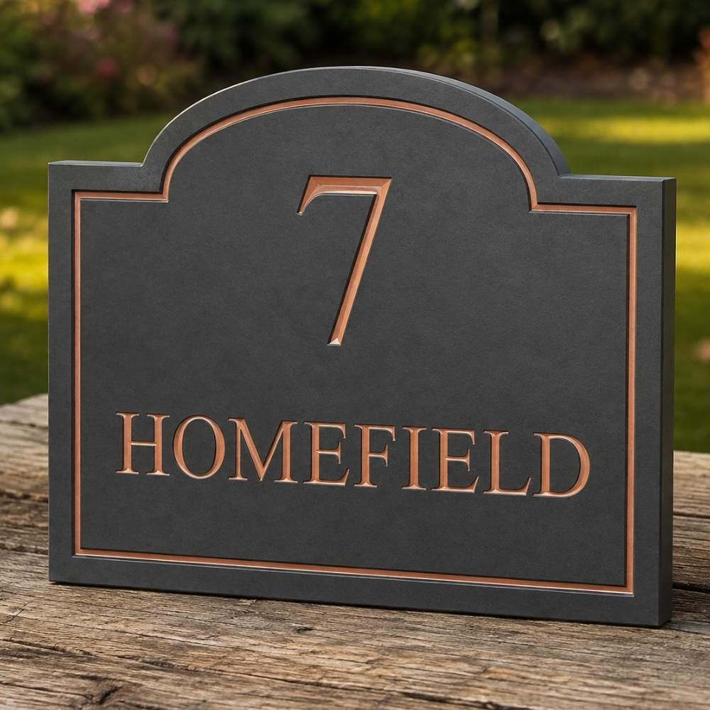 A dark slate house number sign with copper-colored lettering that reads '7 HOMEFIELD', shown resting on a weathered wooden post outdoors with a blurred green garden background.