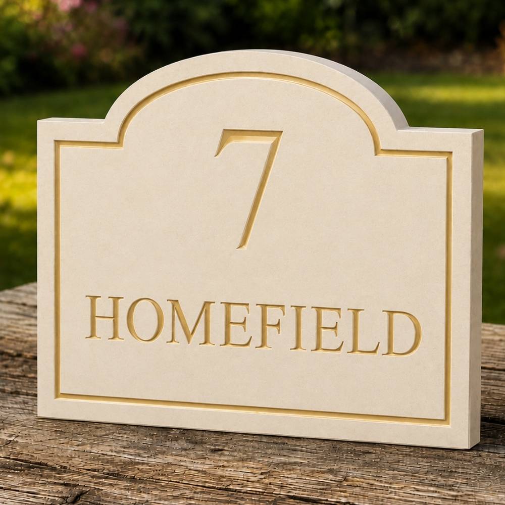 A limestone arched house number plaque with gold-toned lettering reading '7 HOMEFIELD', displayed on a weathered wooden post outdoors with a blurred green garden background.