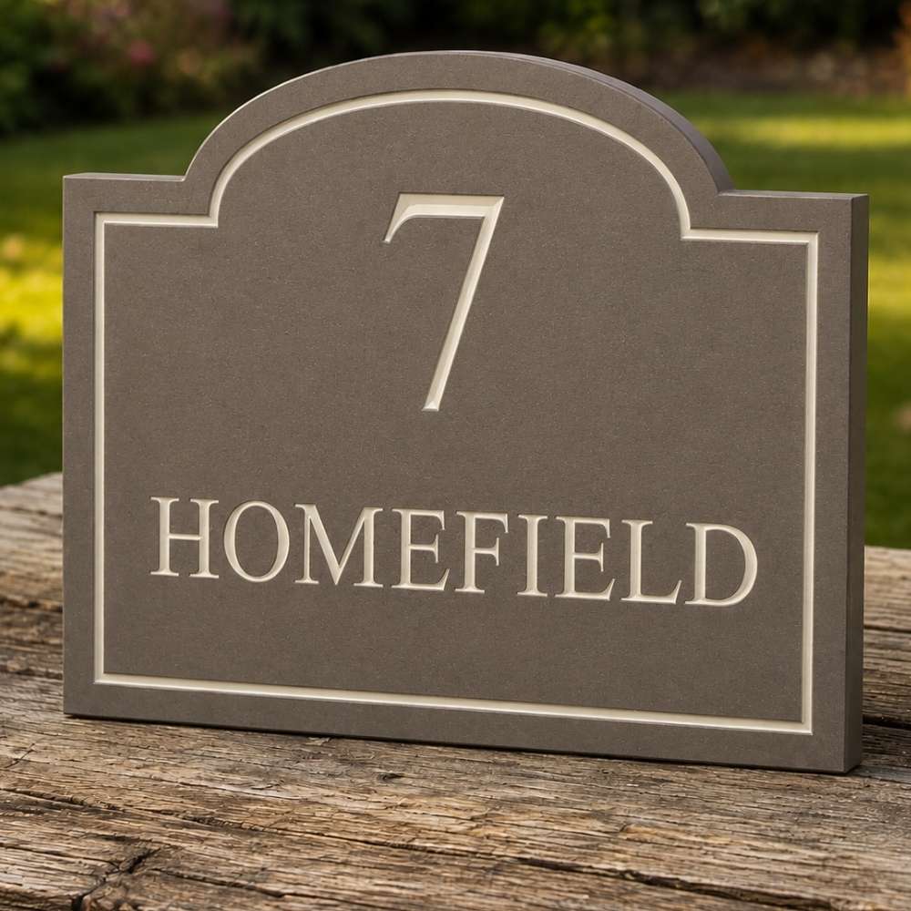 A mink house number plaque reading '7 HOMEFIELD' in raised cream lettering on a dark gray background, shown standing on a weathered wooden surface outdoors with blurred greenery behind it.