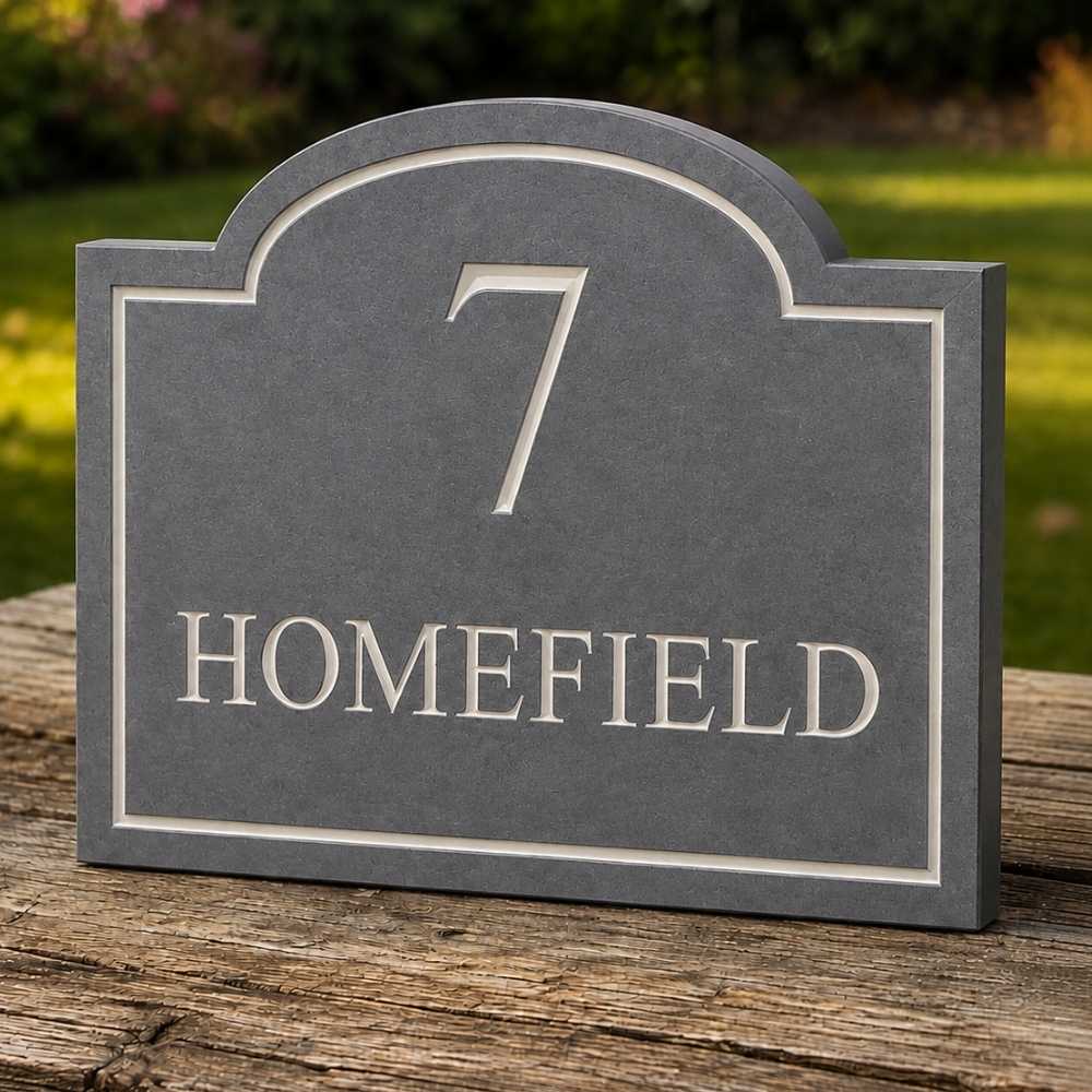 A slate custom house number plaque with the number 7 and the word HOMEFIELD in raised silver lettering, shown standing on a rustic wooden surface with a blurred green garden background.