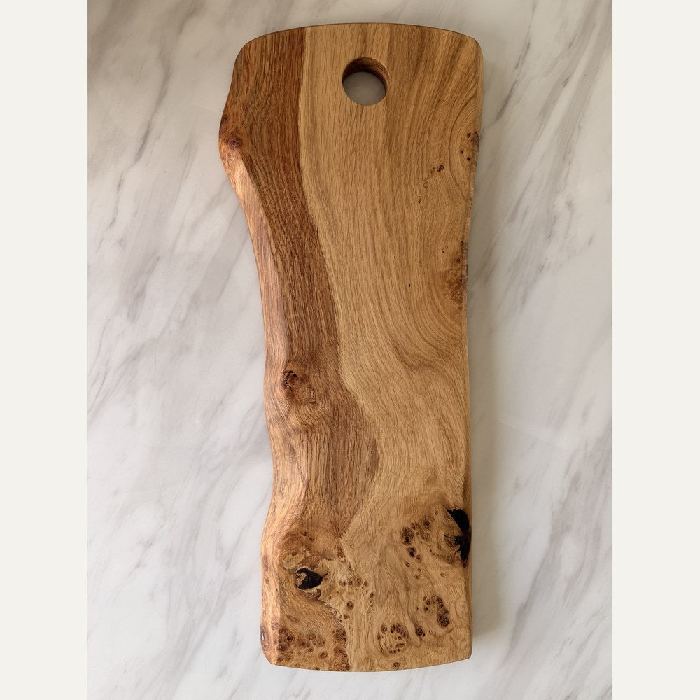 A rectangular wooden serving board with a live edge and a circular hole near the top, photographed from above on a white marble surface.