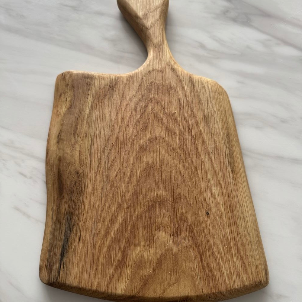 A rectangular wooden serving board with a handle, featuring a natural warm brown wood grain, photographed on a white marble surface.