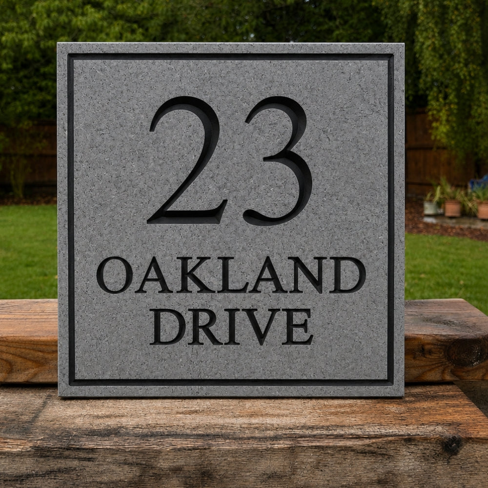 A square granite stone or concrete house number plaque reading “23 OAKLAND DRIVE,” mounted on a wooden post outdoors with grass and trees in the background.