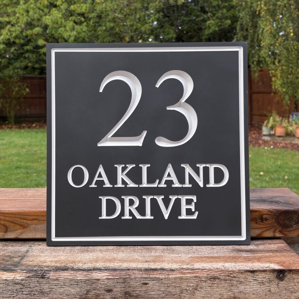 A square dark slate house number sign with silver lettering that reads '23 OAKLAND DRIVE', displayed outdoors on a wooden surface with a garden background.