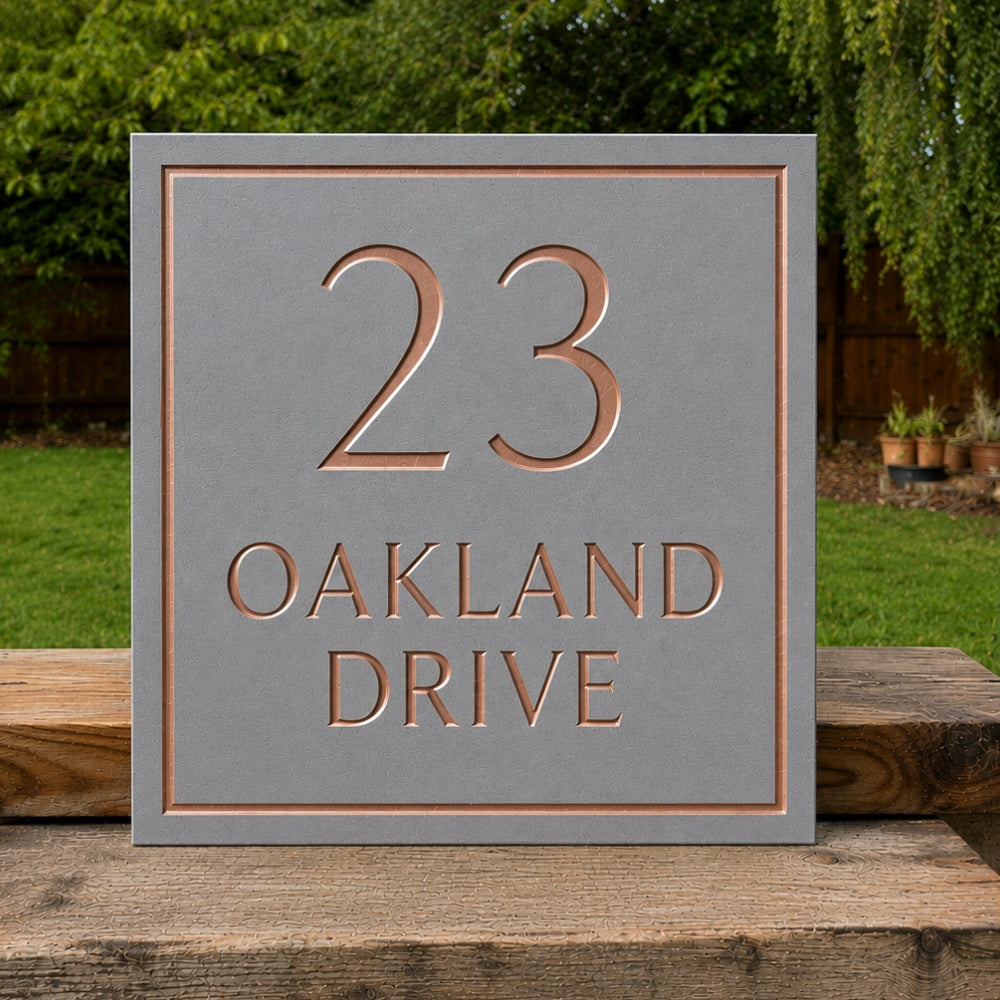 A square grey house number plaque with raised copper-toned lettering reading '23 OAKLAND DRIVE', displayed outdoors on a wooden surface with green grass and trees in the background.