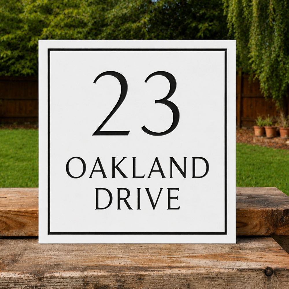 A square white house sign with a black border and the text '23 OAKLAND DRIVE' in black serif font, displayed outdoors on a wooden surface with a blurred green garden background.
