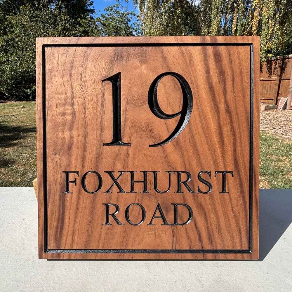 A square walnut wooden house number sign with black lettering reading '19 FOXHURST ROAD', photographed outdoors on a concrete surface with trees and grass in the background.