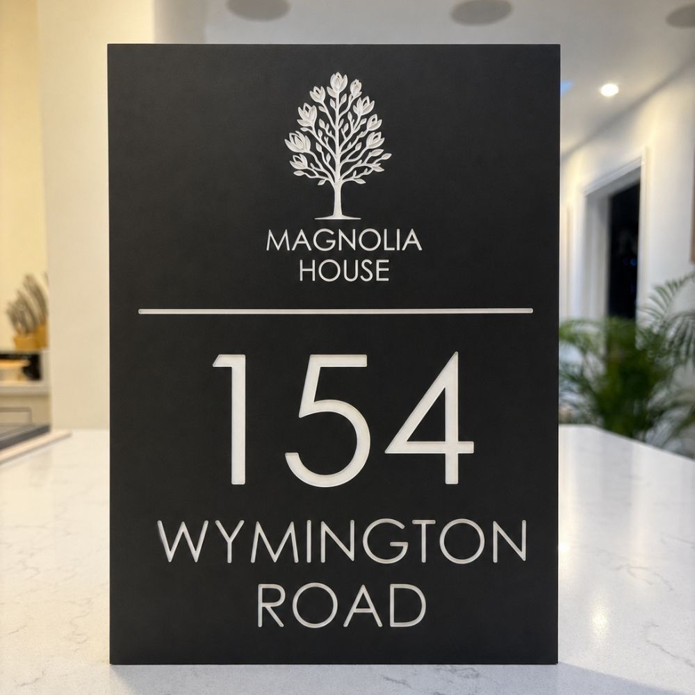 A dark slate house number sign for Magnolia House at 154 Wynington Road, featuring a white tree logo and elegant serif lettering, displayed on a light marble countertop with a blurred modern interior background.
