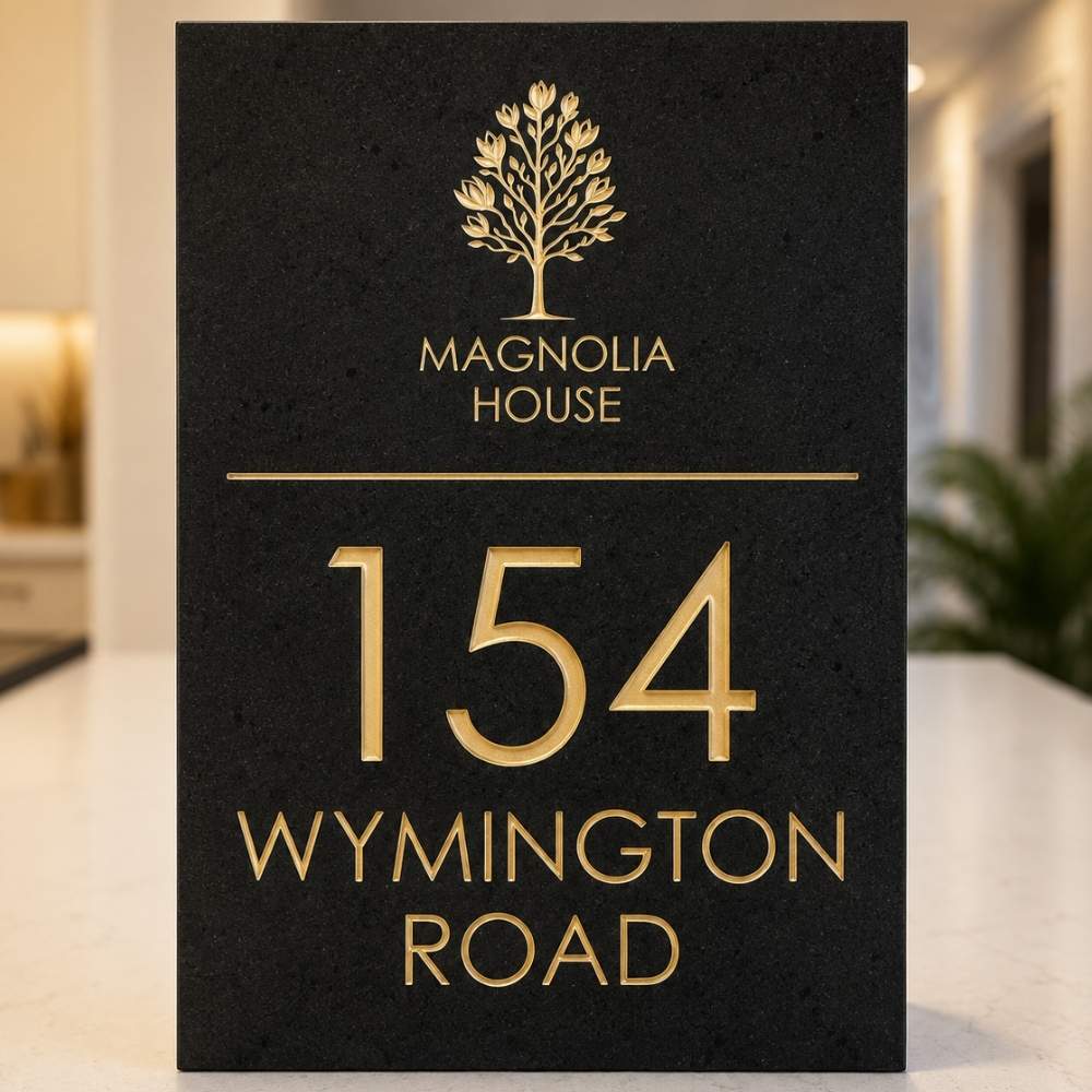 A charcoal rectangular house sign with gold lettering and a gold tree logo, reading 'MAGNOLIA HOUSE', '154', and 'WYMINGTON ROAD', standing on a light surface in an indoor setting.