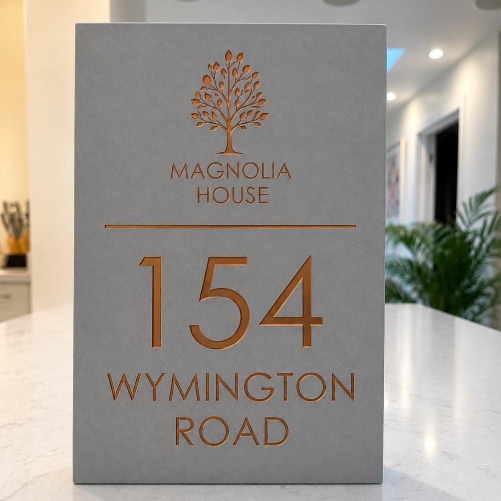 A grey house number sign for Magnolia House at 154 Wynington Road, featuring a metallic copper foil tree logo and lettering, displayed on a white marble counter in a modern interior setting.