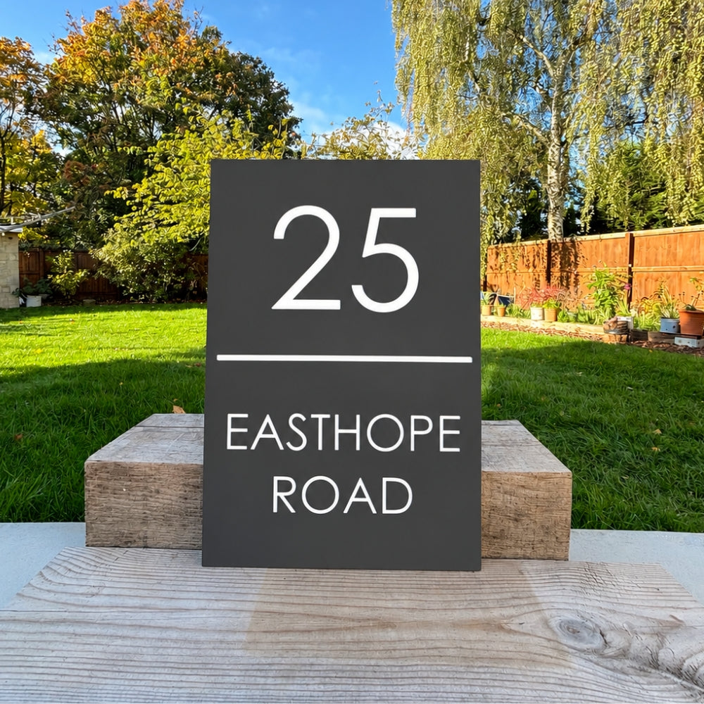 A rectangular dark slate house number sign reading '25 EASTHOPE ROAD' in white text, displayed outdoors on wooden blocks with a grassy backyard and trees in the background.