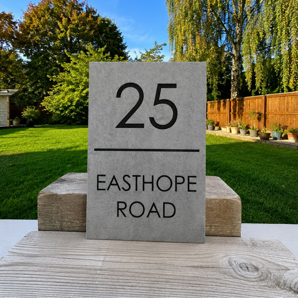 A rectangular granite house number plaque showing '25 EASTHOPE ROAD' in black text, displayed outdoors on a wooden surface with a green lawn and trees in the background.