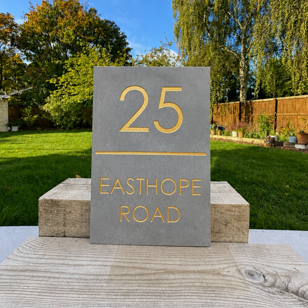 A rectangular grey house number plaque with gold lettering reading '25 EASTHOPE ROAD' is displayed outdoors on two wooden blocks in a garden setting.