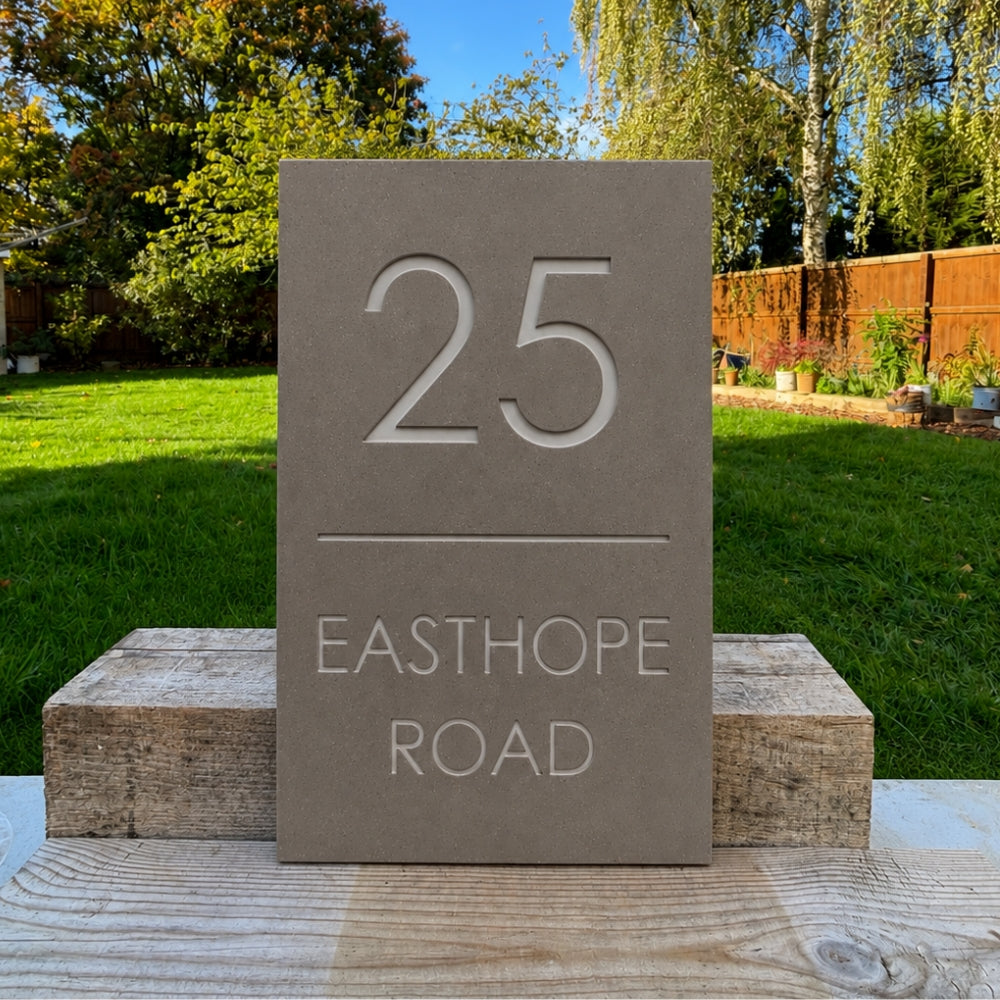 A rectangular mink house number plaque displaying '25 EASTHOPE ROAD' in raised white lettering, mounted on two wooden blocks outdoors with a green lawn and trees in the background.