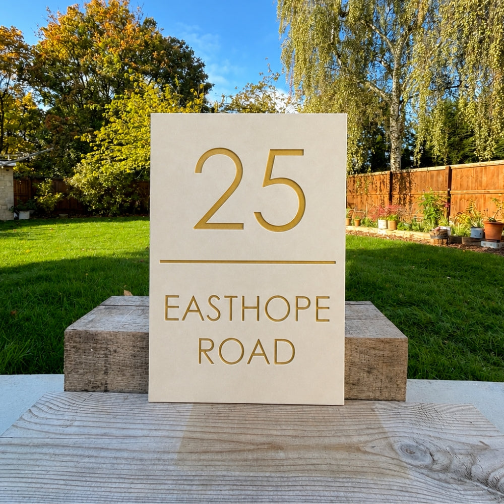 A rectangular quartz outdoor house number sign reading '25 EASTHOPE ROAD' in gold lettering, displayed on wooden blocks in a backyard setting.