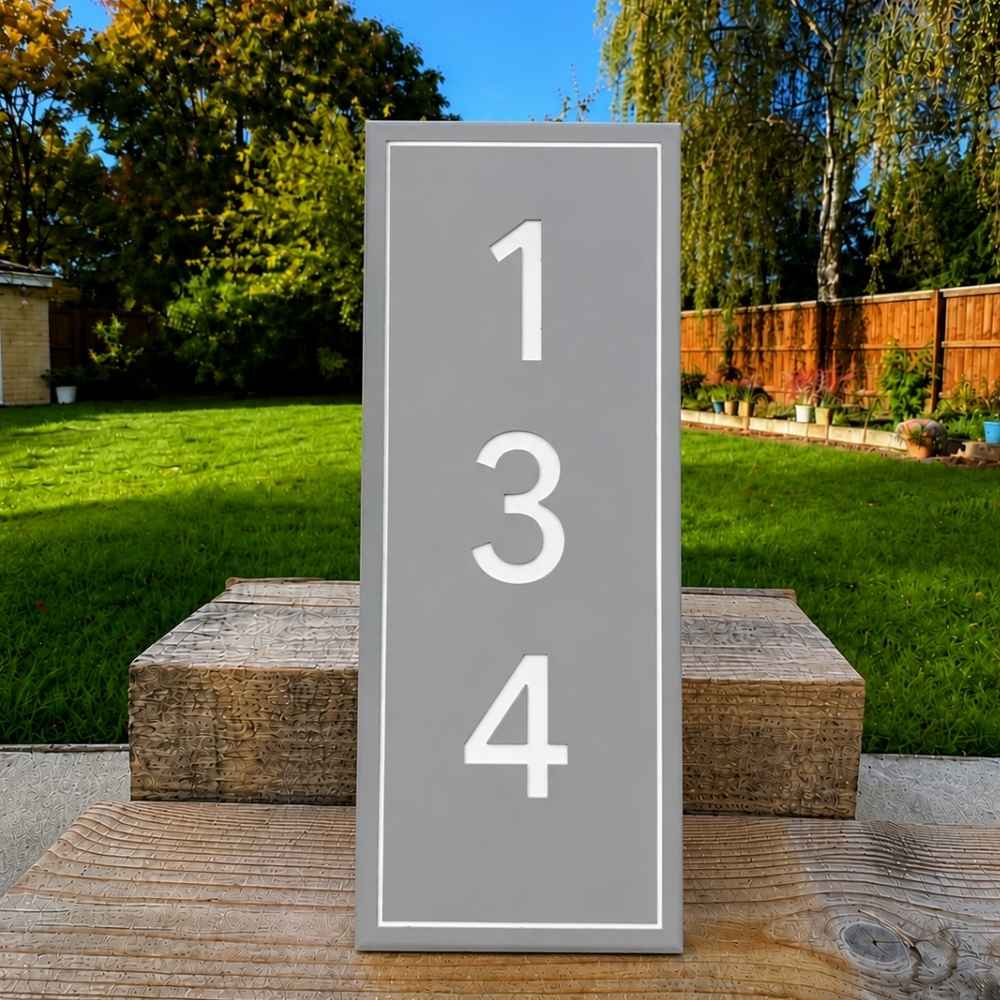 A vertical gray house number plaque with white numerals '134' and a thin white border, displayed outdoors on a wooden post in a grassy backyard.